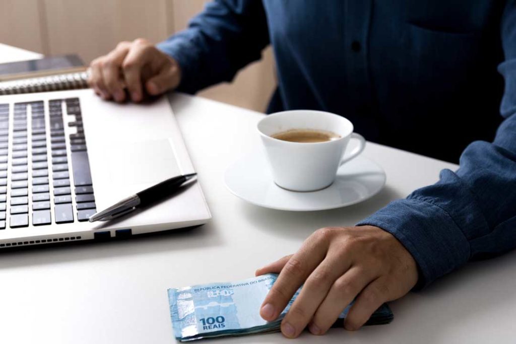 A imagem tem como foco, a mão de uma pessoa, que está a frente de um notebook, segurando cédulas de reais, com um café na mesa
